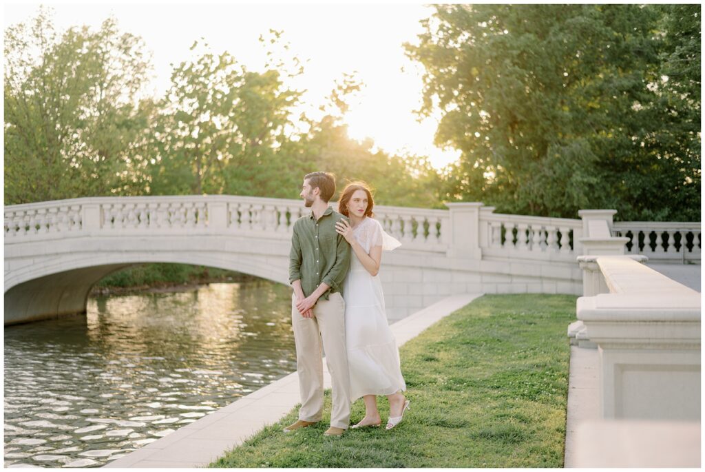 Couple standing by a stone bridge over water at sunset in Forest Park, St. Louis, with soft golden light filtering through trees