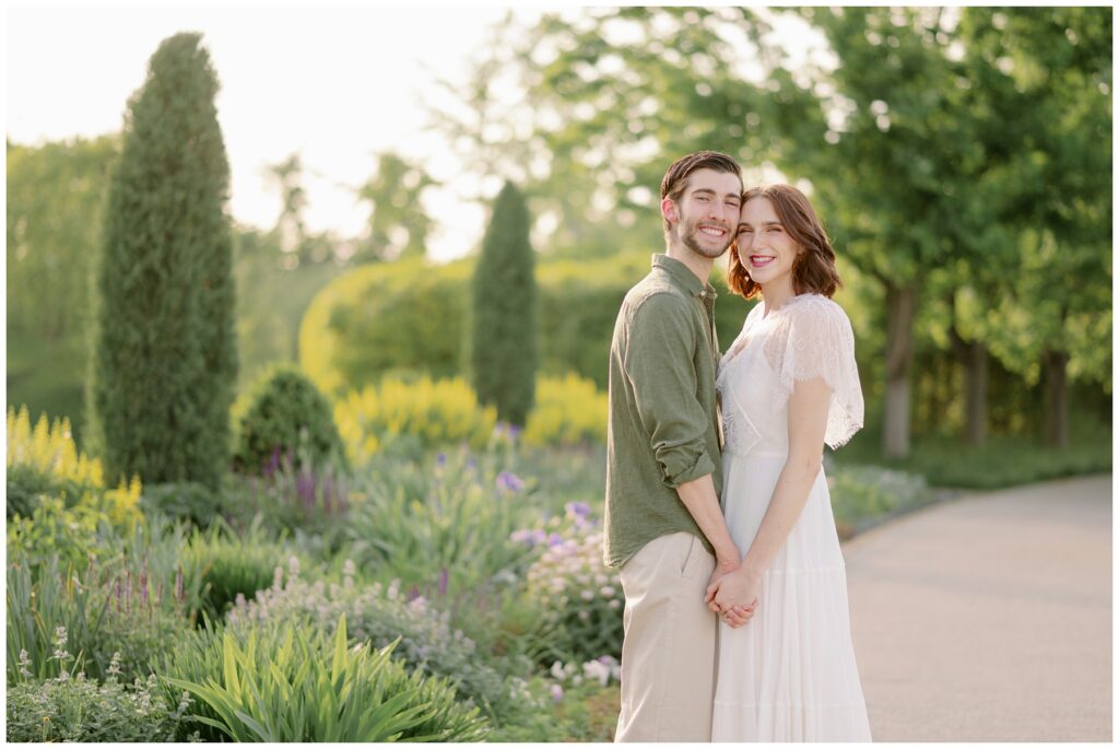 Engaged couple smiling at the camera while holding hands in a lush garden at sunset in Forest Park