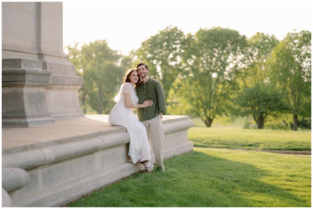Engaged couple sitting together on a stone ledge, smiling at the camera in soft golden light at Forest Park