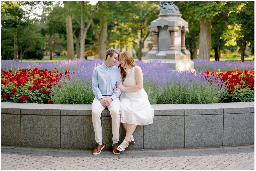 Couple sitting among purple lavender and red flowers in God Quad during summer University of Notre Dame engagement photos.