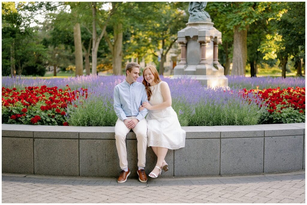 Couple sitting together in front of colorful flowers in God Quad during summer University of Notre Dame engagement photos.