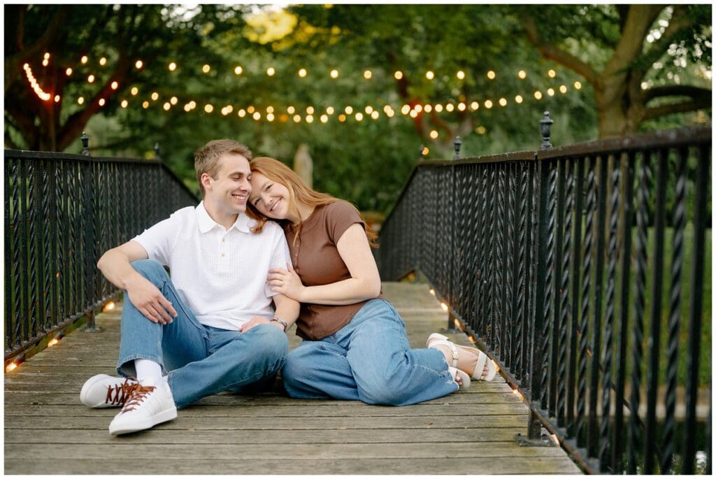 Couple sitting together on the illuminated bridge during summer Saint Mary's College engagement photos at sunset.
