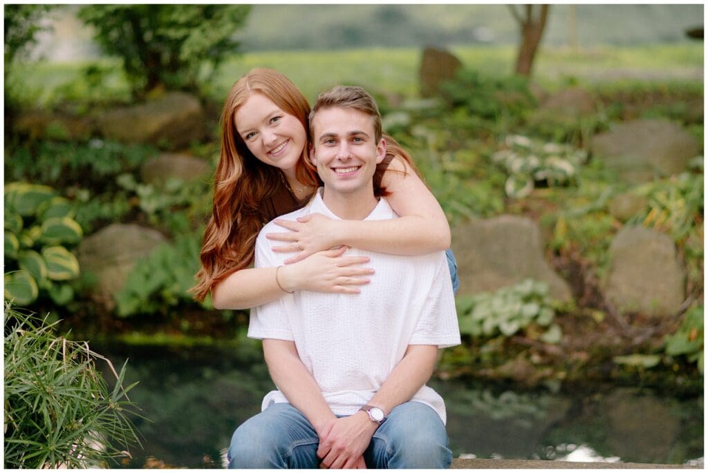 Natural engagement portrait of the smiling couple embracing beside a peaceful garden pond with lush greenery in the background.